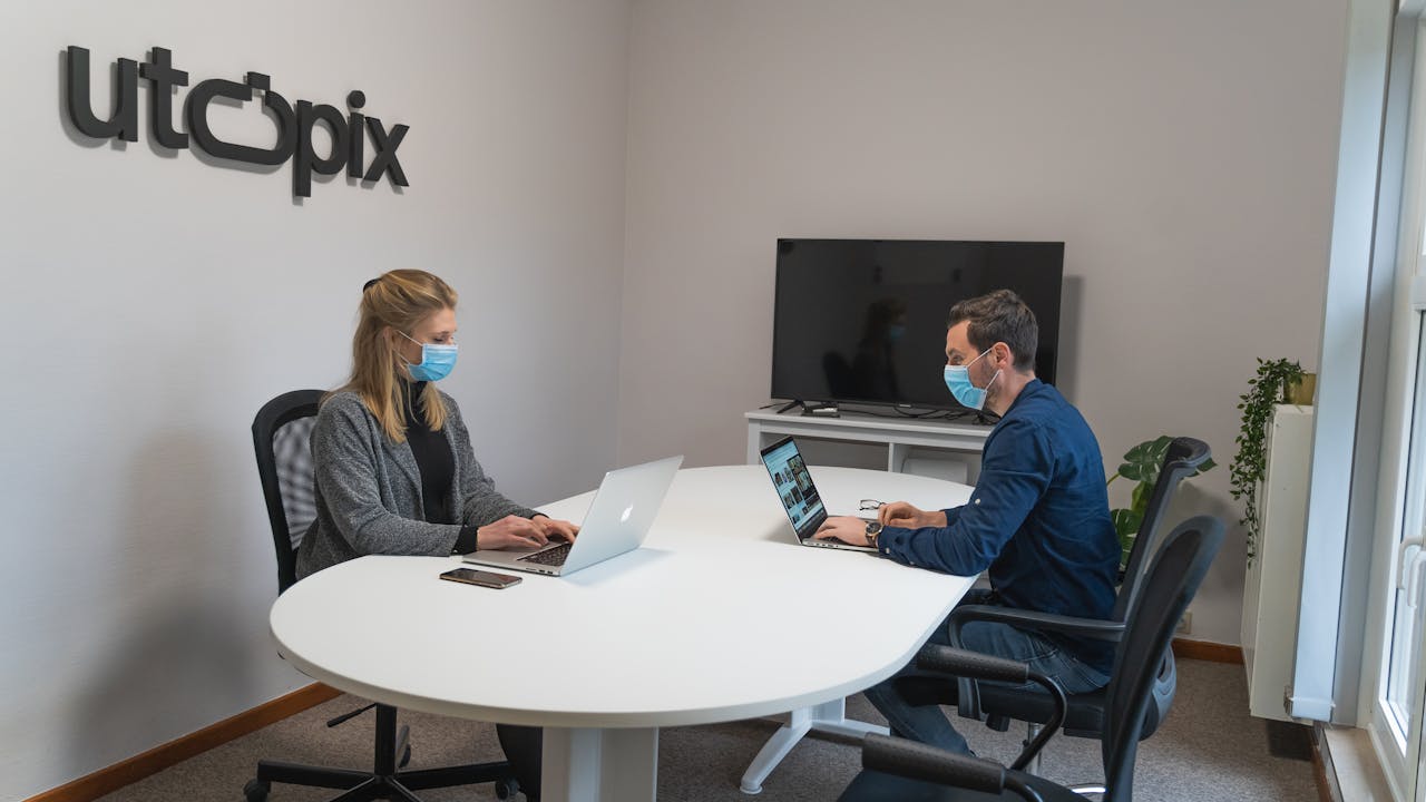 Two coworkers in an office setting wearing masks, maintaining social distancing while working on laptops.